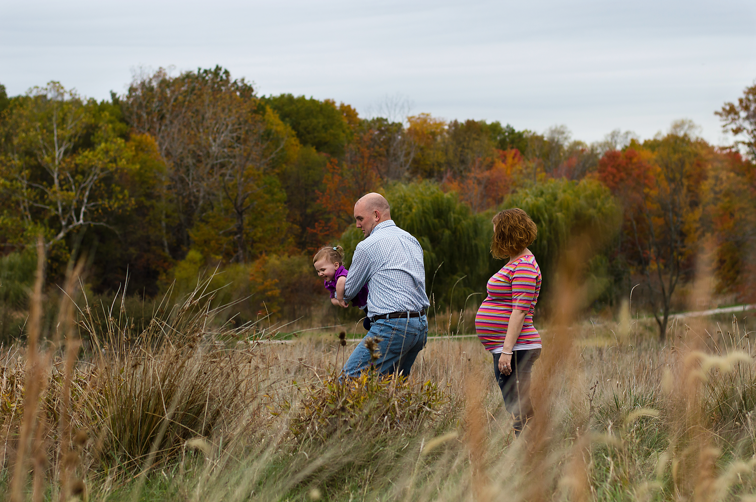 outdoor-maternity-photography-west-creek-reservation-family-fun-at-the-photoshoot-dad-spinning-his-little-girl-with-pregnant-mom-watches-them.png