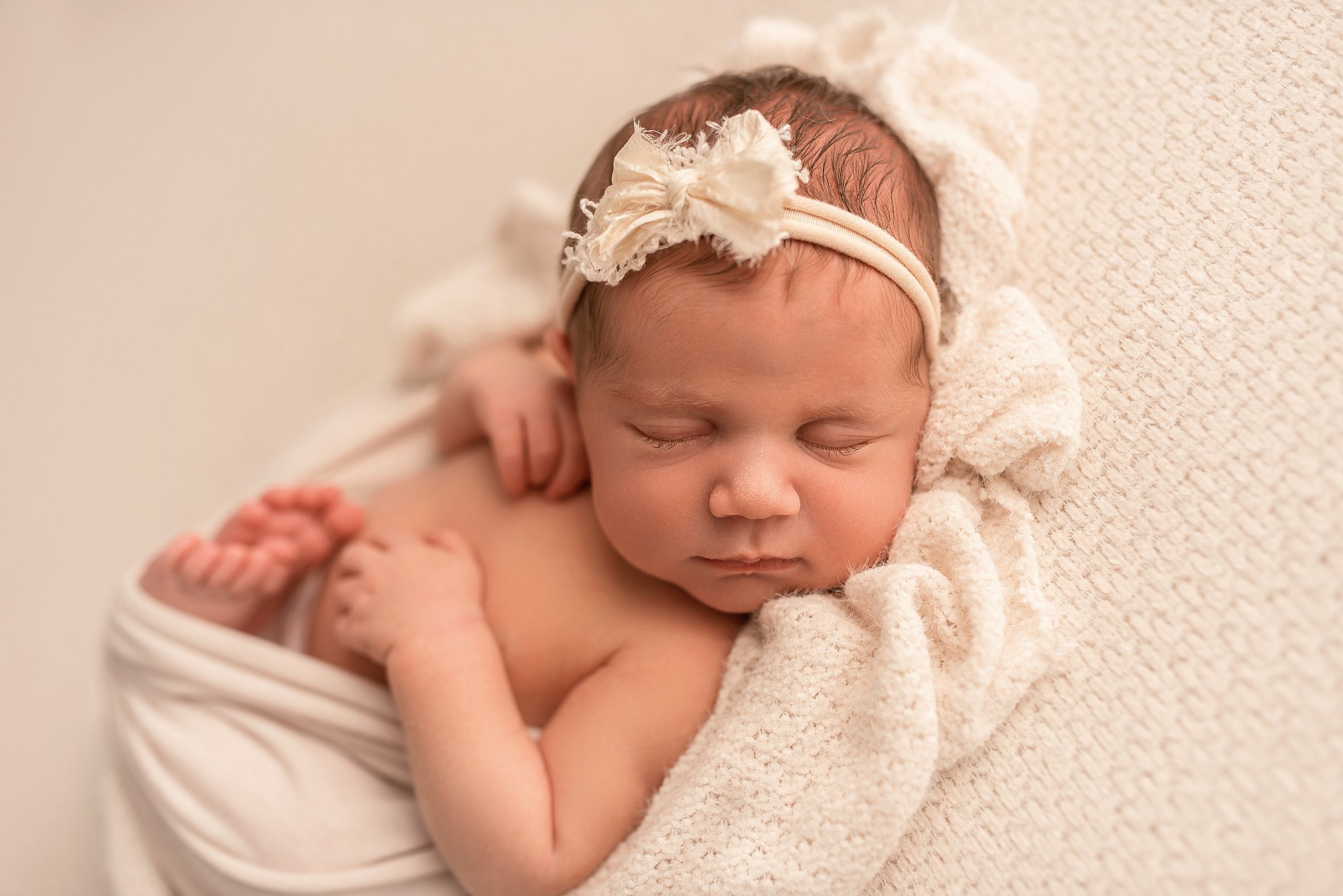 A newborn baby sleeps in a white blanket with a white headband and bow The nanny connection