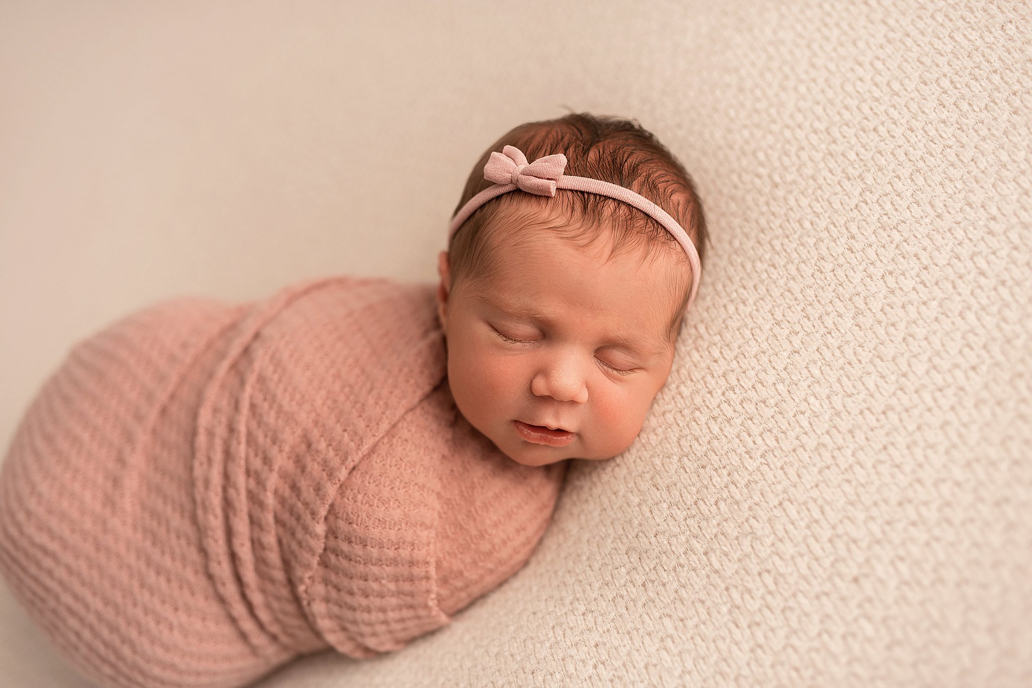 A newborn baby sleeps on a knit pad in a pink swaddle blanket