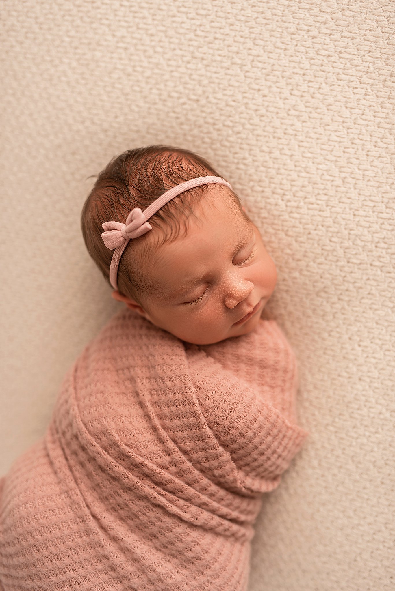 A newborn baby sleeps while swaddled in a pink blanket and pink headband with a bow The nanny connection