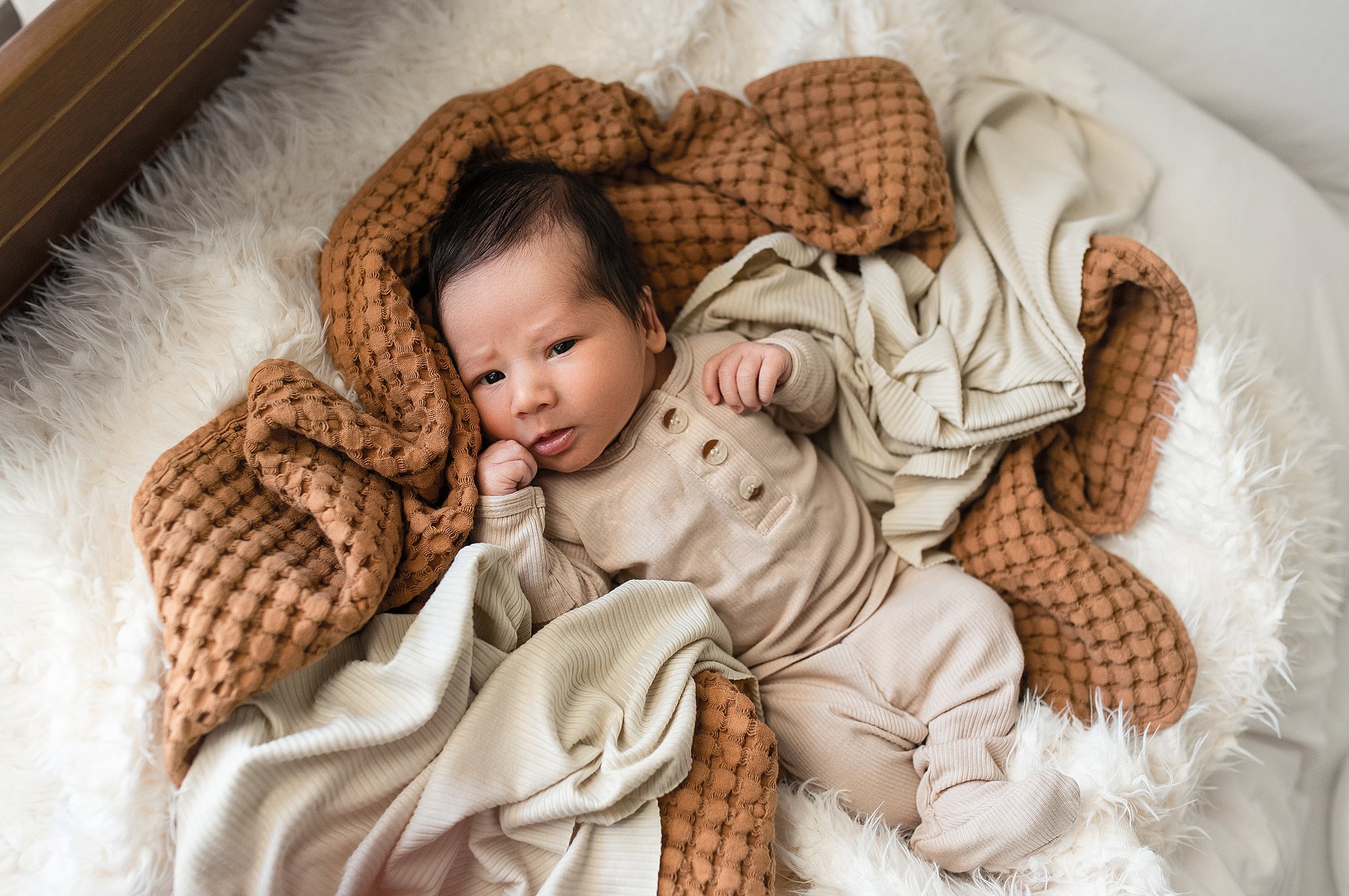 A newborn baby lays on a bed of blankets in a beige onesie with eyes open