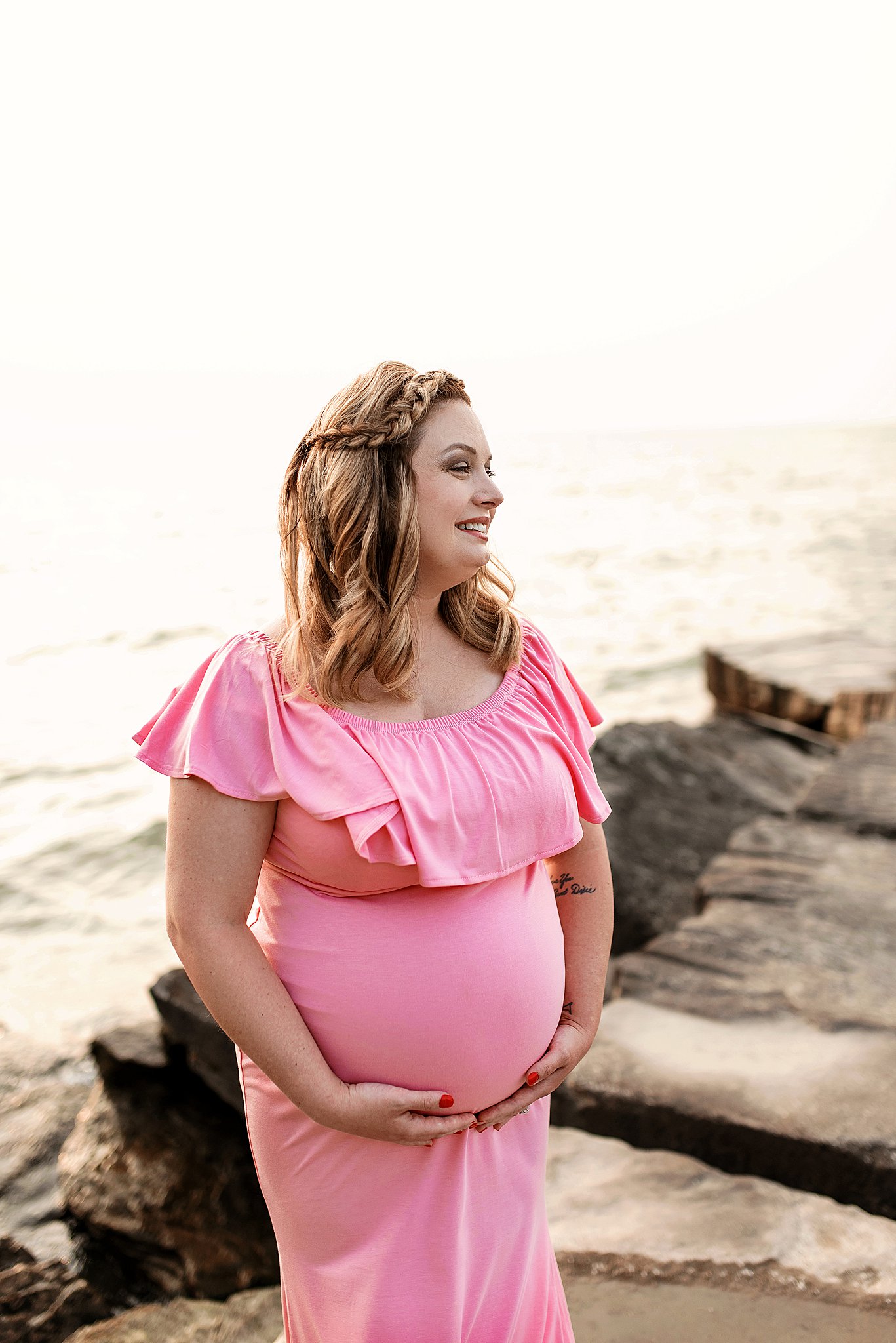 A mother to be in a pink dress stands on some rocks on the beach holding her bump birth center cleveland ohio