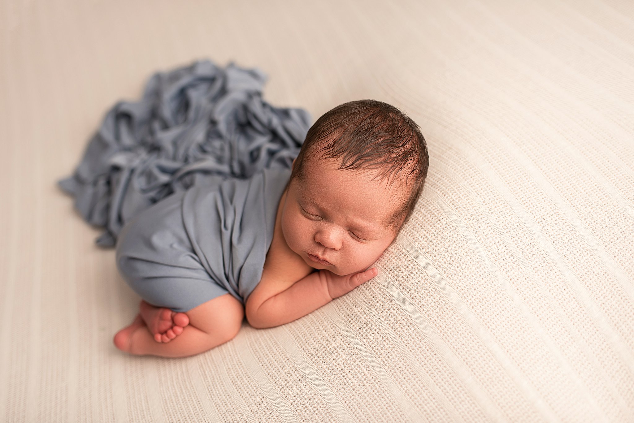 A newborn baby sleeps in froggy pose while wrapped in a blue blanket
