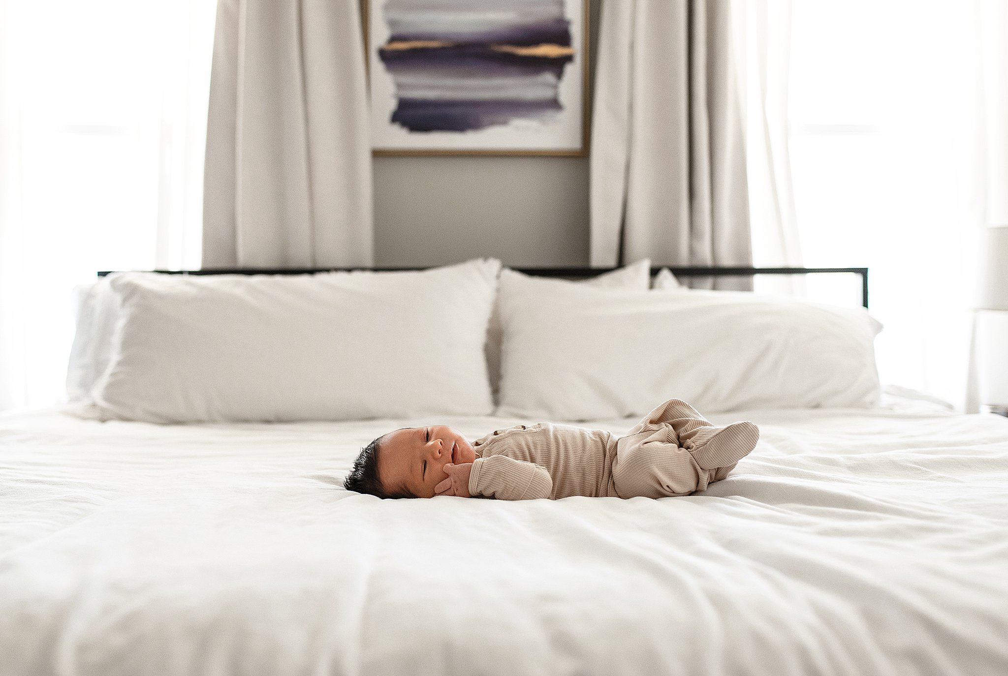 A newborn baby lays on a white bed in front of windows in a beige onesie