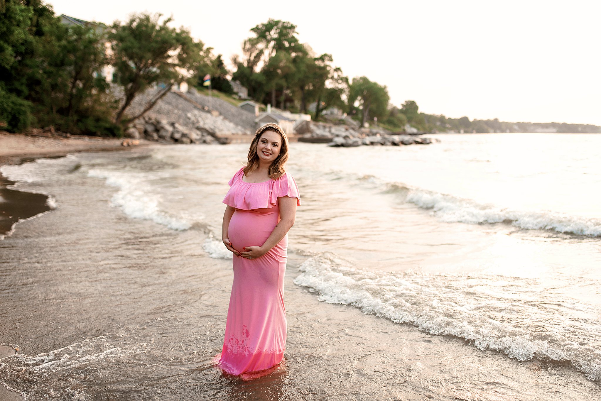 A mother to be in a bright pink maternity gown stands in the water on a beach holding her bump midwives in cleveland ohio