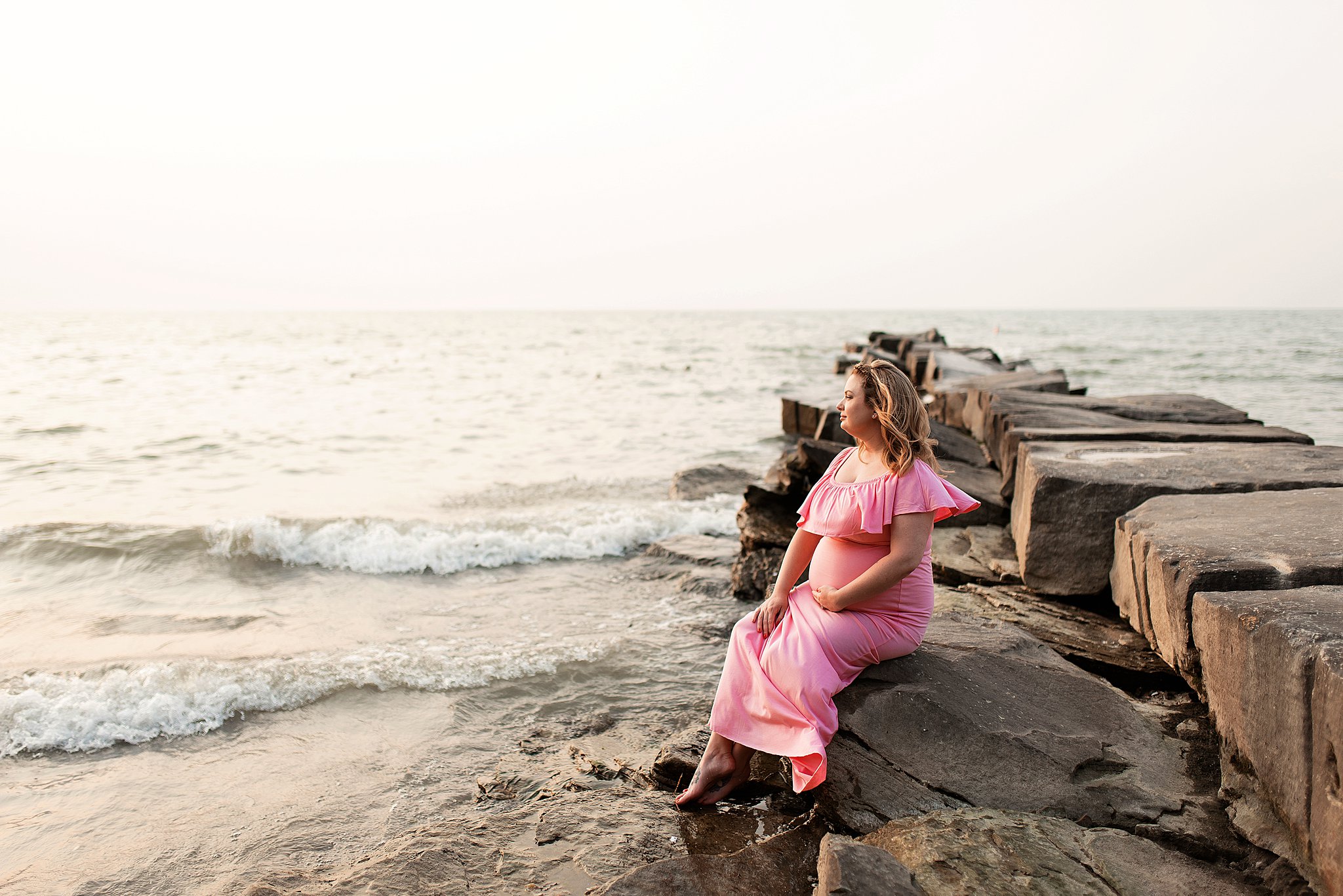 A mom to be sits on some rocks at sunset with her feet dangling in the ocean
