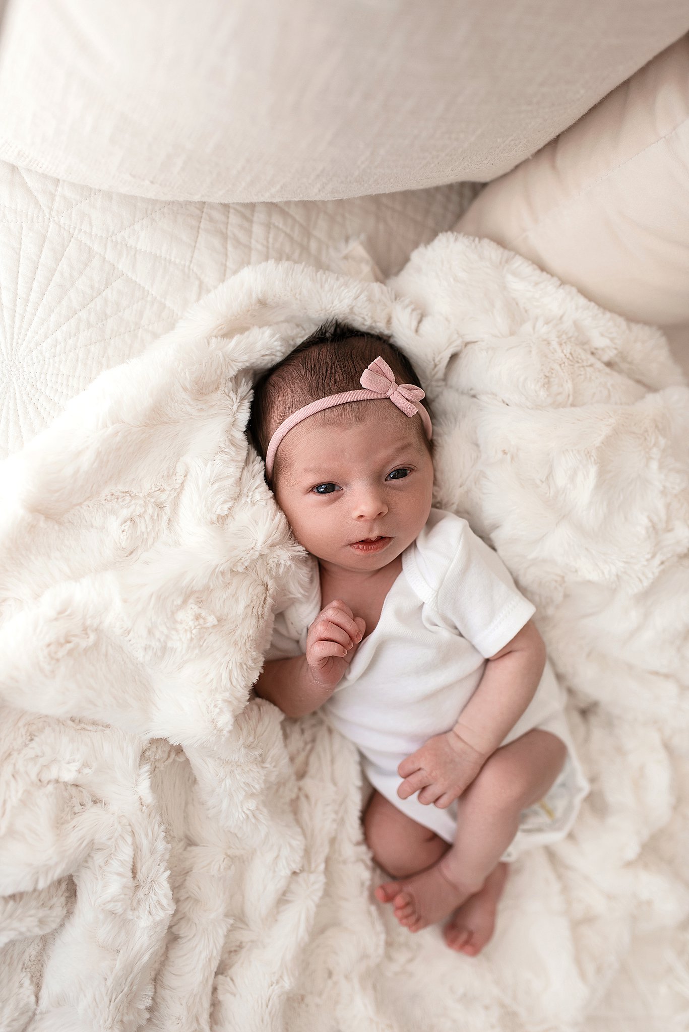 A newborn baby lays on a white furry blanket on a couch with eyes open and a pink bow headband